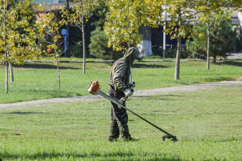 A man in a green jacket operates a lawn mower on a grassy area, focused on maintaining the lawn's appearance.