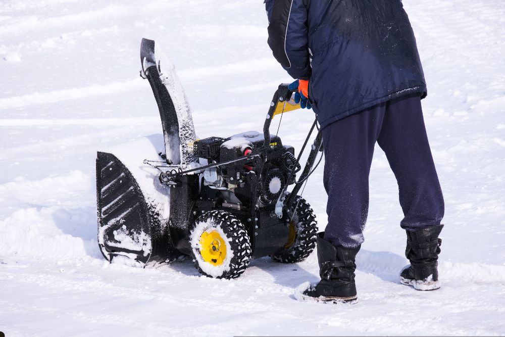 A man operates a snow blower, clearing a path through freshly fallen snow in a residential area.